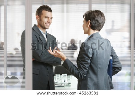 Businessman and businesswoman leaving meeting room, talking at the door, smiling.