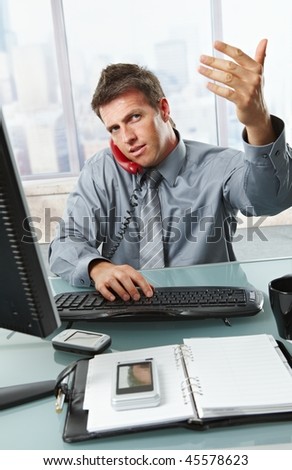 Mid-adult businessman on landline call waving someone out of picture to enter while busy working with computer in office.