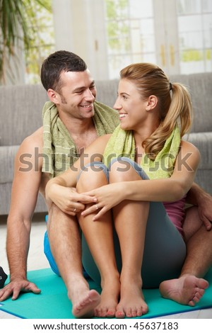 Happy couple resting after training, sitting on fitness mat in living room, looking at each other smiling.