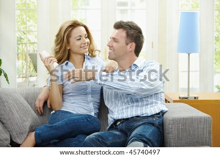 Young couple watching TV at home, sitting on couch, holding remote control in hand.