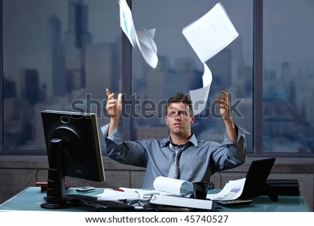 Exhausted professinal throwing documents into air sitting at office desk in overtime.