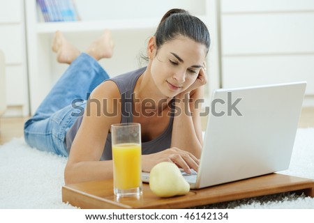 Happy woman lying on floor at home and working on laptop computer, smiling.