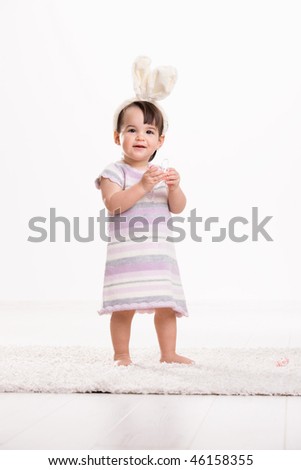 Happy baby girl in easter costume standing on carpet, playing with easter eggs, smiling. Isolated on white background.