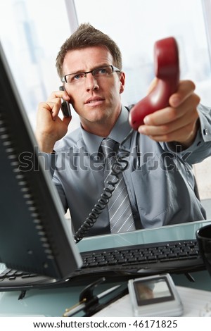 Businessman with glasses busy talking on mobile phone handing over landline call to answer in office.