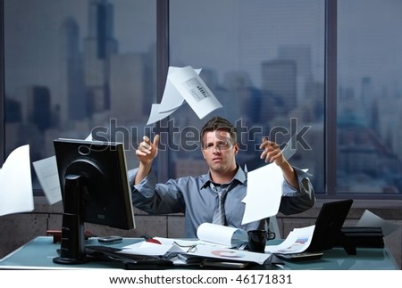 Exhausted businessman throwing documents into air sitting at office desk in overtime.