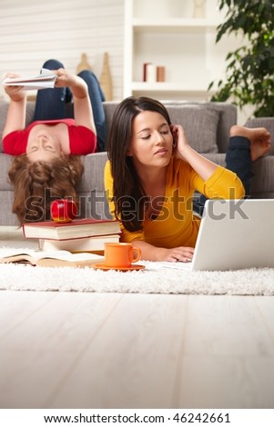 Teenage girls studying at home in living room lying on sofa and floor with books and laptop.