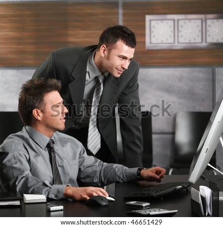 Two businessmen working together with computer at office desk, looking at screen.