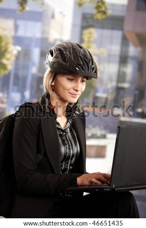 Portrait of young businesswoman wearing bike helmet, sitting in front of office building, using laptop computer.