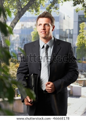Smart executive businessman in suit standing on office building courtyard holding briefcase.