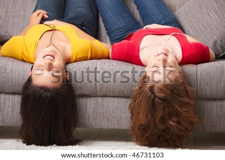 Teen girls lying on couch upside down, listening to music in earbuds, eyes closed.