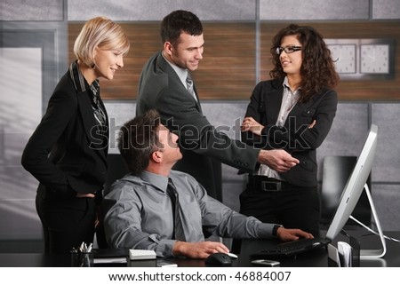 Businessman sitting at office desk, looking up at happy colleagues chatting.