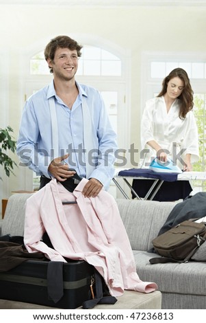 Young businessman preparing for business trip. Woman ironing in the background.