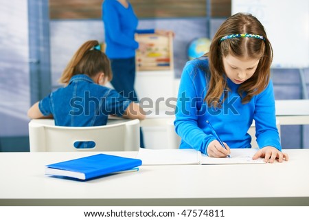 Elementary age schoolgirls writing tests at class seated aside, teacher standing in the background.