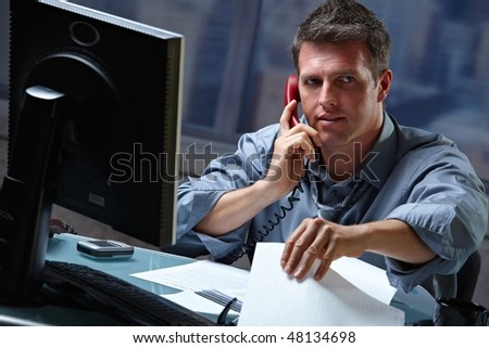 Mid-adult businessman speaking on landline phone working overtime in office checking papers.