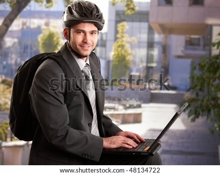 Portrait of young businessman wearing bike helmet, sitting in front of office building, using laptop computer.