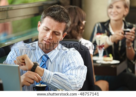 Businessman sitting at table in cafe, waiting for somebody looking at his watch. Young women having sweets in the background.