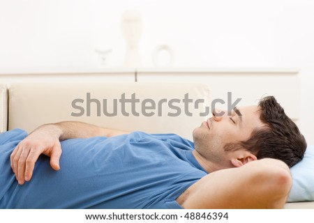Young man in blue t-shirt sleeping on couch in living room.