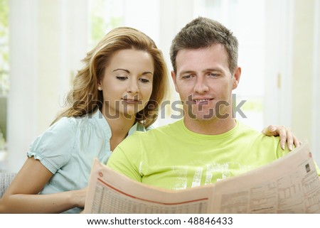 Love couple reading newspaper together on couch at home, smiling.
