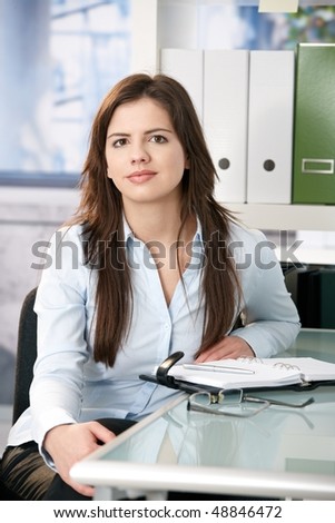 Attractive woman sitting at office desk with organizer, looking at camera, smiling.