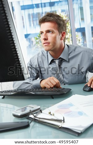 Mid-adult businessman concentrating on computer work at office desk looking at screen.