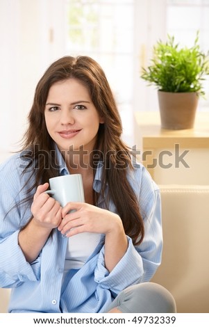 Pretty young woman sitting on couch at home in morning sunlight holding mug, looking at camera, smiling.