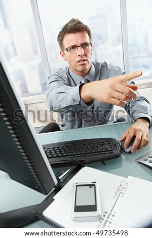 Serious businessman talking on landline phone gesturing out of picture sitting at office desk.