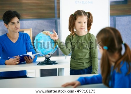 Young schoolgirl pointing at globe in class, looking at another pupil, teacher smiling.