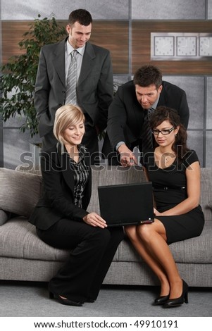 Young businesswomen sitting at couch in office holding laptop computer, businessman showing someting on screen.