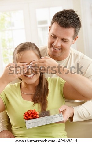 Smiling man giving surprise, smiling woman covering eyes with hands at home.