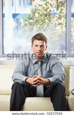 Confident businessman looking determined into camera with folded hands sitting on leather couch in sunny background.