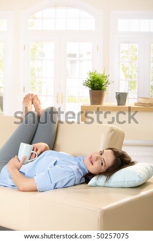 Young woman lying on sofa with feet up holding coffee mug in hand, smiling.