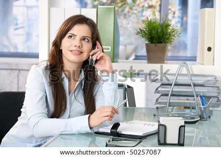 Female assistant sitting in office, speaking on mobile phone, looking up, smiling, holding pen.