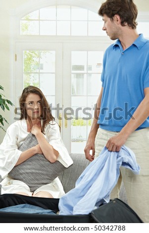 Unhappy couple breaking. Man packing his clothes into suitcase, crying woman hugging pillow in the background. Selective focus on woman.