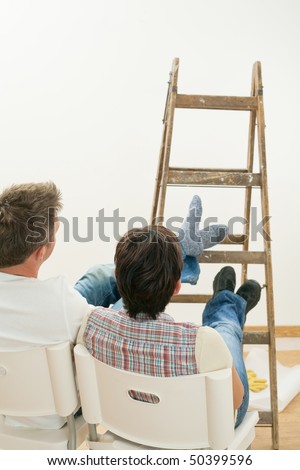Couple relaxing after work, looking at freshly painted white wall, sitting with feet up on ladder.
