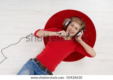 Woman lying on round red carpet of living room floor with headphones holding microphone, singing in high angle view.