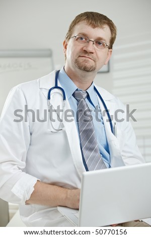 Medical office - male doctor sitting on desk holding laptop computer in hand, looking away, thinking.