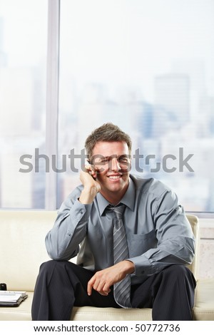 Happy businessman calling on mobile phone sitting on leather couch in office smiling.