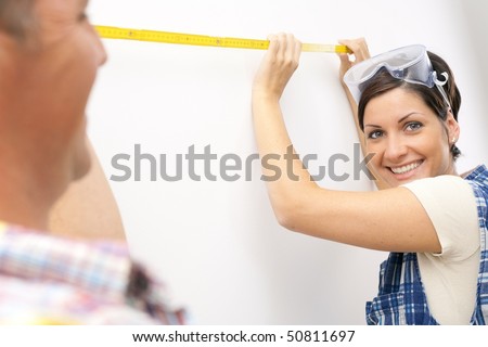 Young woman smiling at camera holding measuring stick in focus, man laughing.