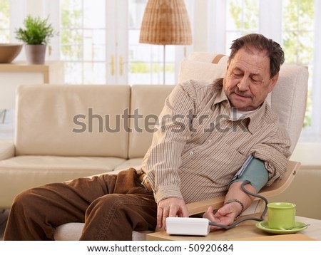 Old man sitting in armchair at home, measuring his blood pressure, smiling.