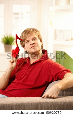 Young man holding red female slipper in hand sitting on couch looking affectionate.