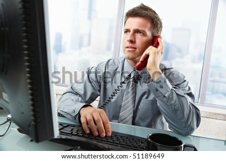 Determined businessman discussing computer work on landline phone while looking at screen typing on keyboard at office desk.