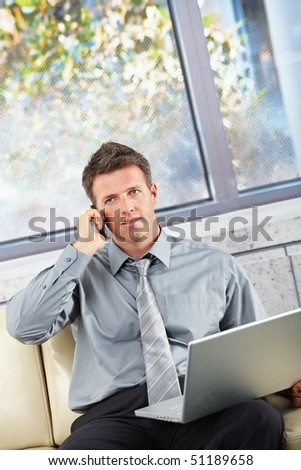 Businessman on cell phone sitting on a beige sofa with computer in lap.