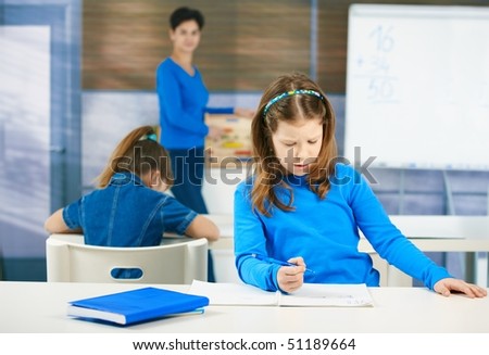 Schoolgirl thinking while filling out test at class, teacher standing in background.