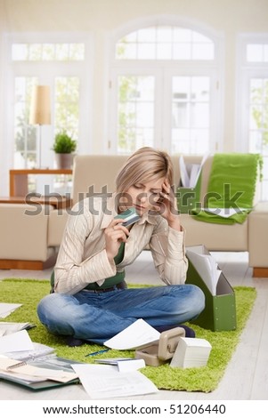 Troubled woman sitting on floor with crossed legs, looking at documents holding credit card in living room.