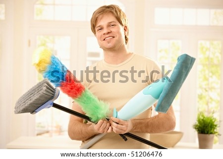 Young man smiling confidently with cleaning tools, holding mop, broom and flannel.