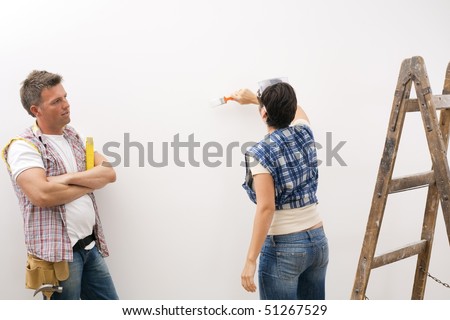Woman painting white wall, man focusing on work standing with folded arms.