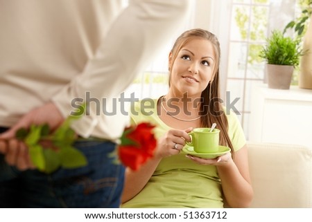 Man bringing flower to woman sitting on couch at home with tea cup, smiling.