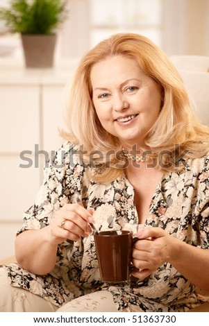Happy senior woman stirring coffee with spoon, holding coffee mug, laughing at camera, sitting in living room.