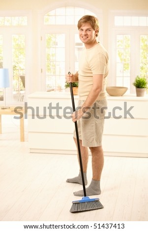 Smiling guy sweeping the floor in living room, looking at camera, standing.