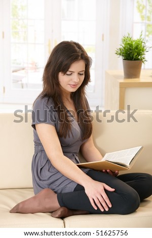Pretty girl sitting on couch, reading book with legs pulled up, smiling.